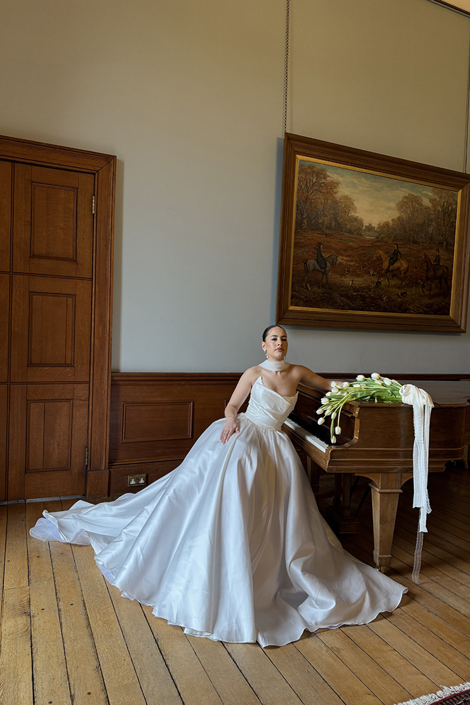 Bride in a full satin wedding dress poses beside a grand piano with a bouquet of white tulips at a historic Scottish venue.