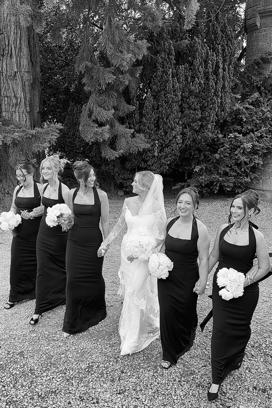 Black-and-white photo of a bride walking outdoors with bridesmaids in matching black halterneck dresses.