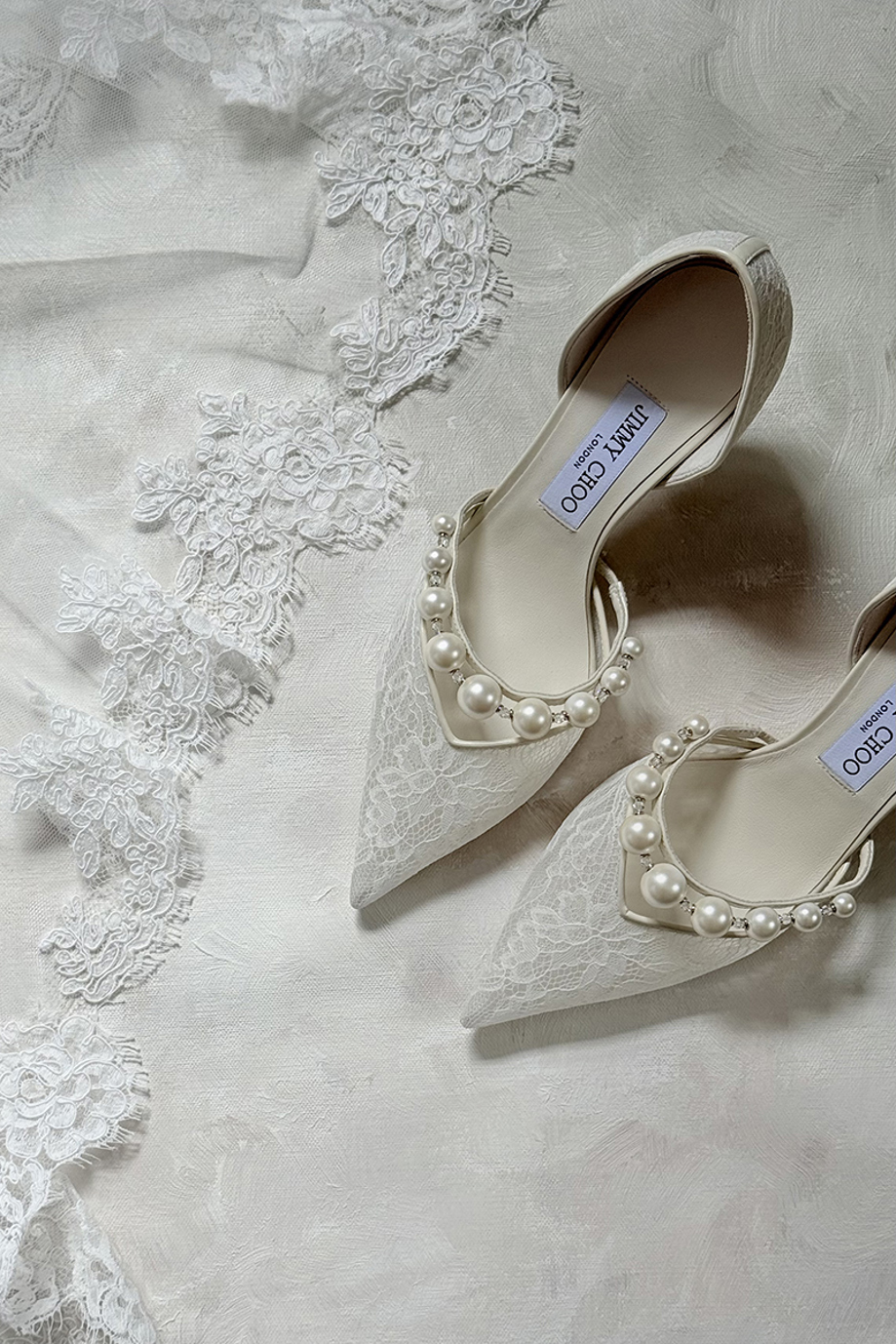 White lace Jimmy Choo bridal shoes decorated with pearls displayed beside an embroidered wedding veil.