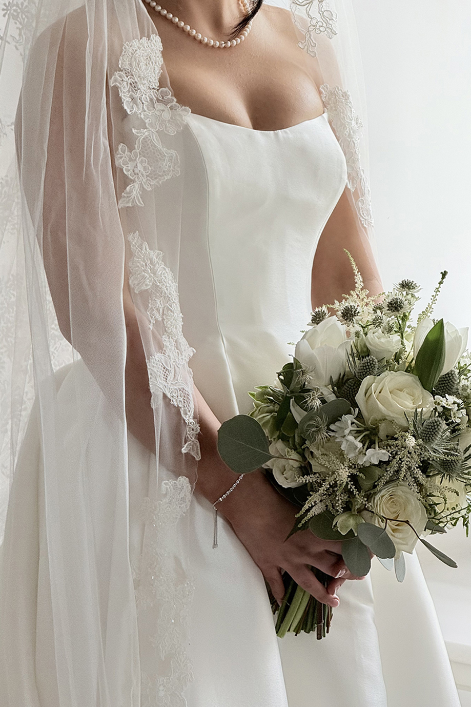 Close-up of a bride holding a white and green wedding bouquet beneath a floral lace veil.