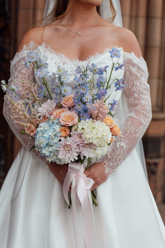 Close-up of a pastel bridal bouquet with blue hydrangeas, peach roses, pink chrysanthemums and delphiniums tied with ribbon.