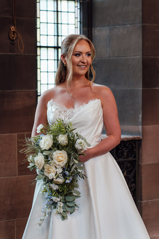 Bride in a satin wedding dress holds a cascading bouquet of white roses, blue thistles and greenery by Maison Bloom Florals.
