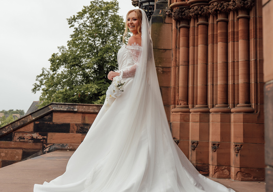 Bride poses outside a Gothic church holding a delicate white orchid wedding bouquet by Maison Bloom Florals.