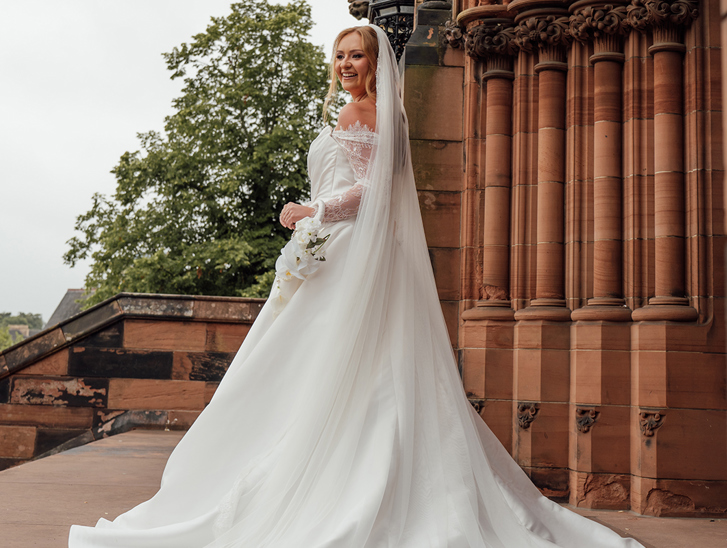 Bride poses outside a Gothic church holding a delicate white orchid wedding bouquet by Maison Bloom Florals.