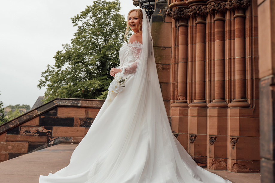 Bride poses outside a Gothic church holding a delicate white orchid wedding bouquet by Maison Bloom Florals.