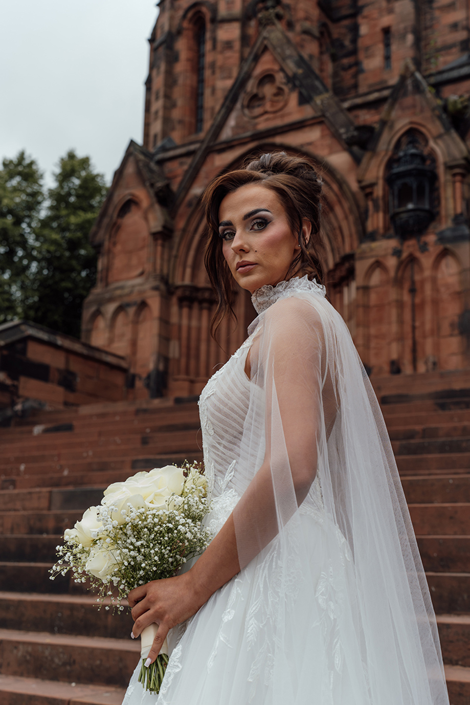 Bride in a lace wedding dress holds a white rose and gypsophila bouquet outside a historic red sandstone church.
