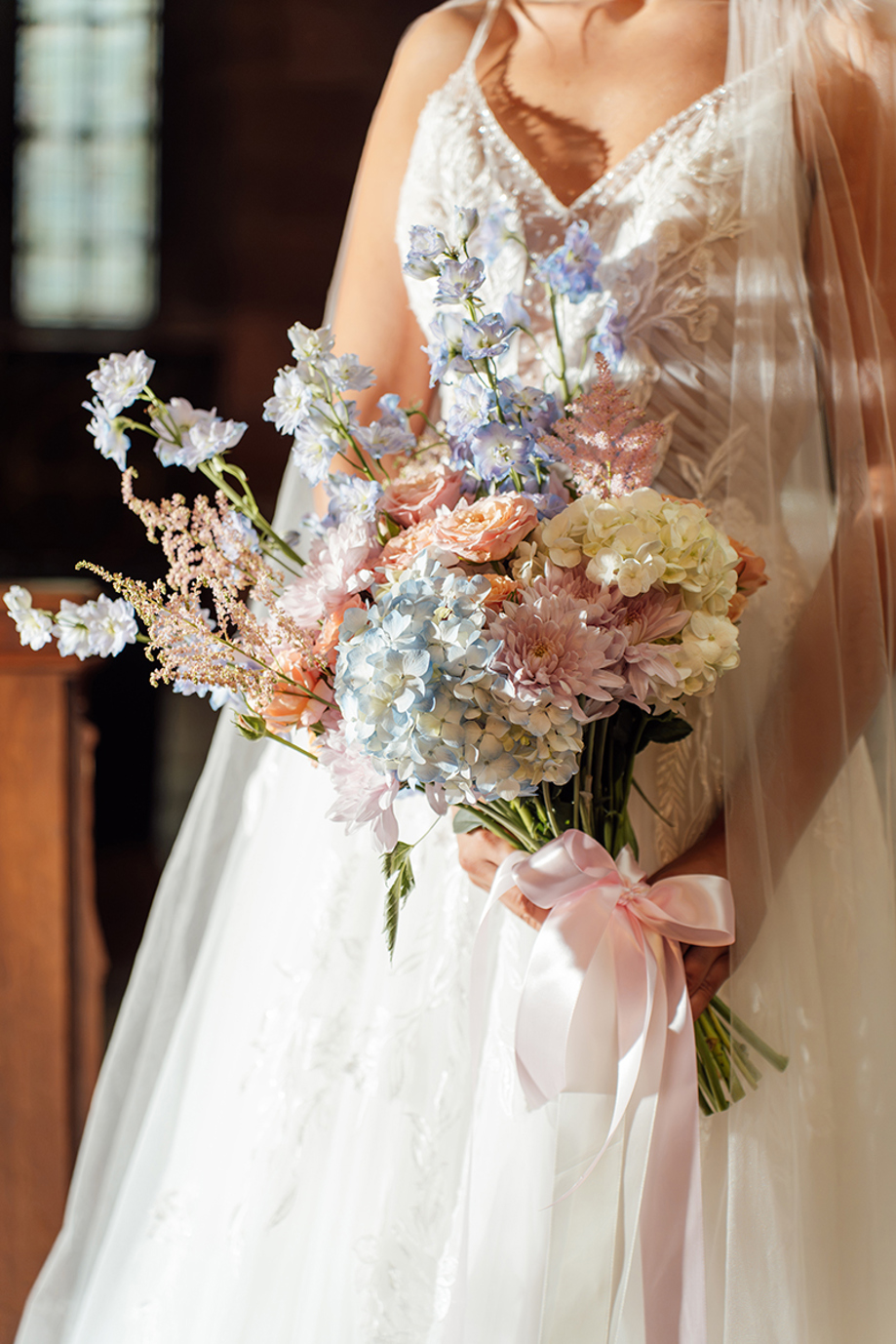 Bride holds a pastel wedding bouquet of blue hydrangeas, peach roses, pink flowers and delphiniums tied with ribbon.