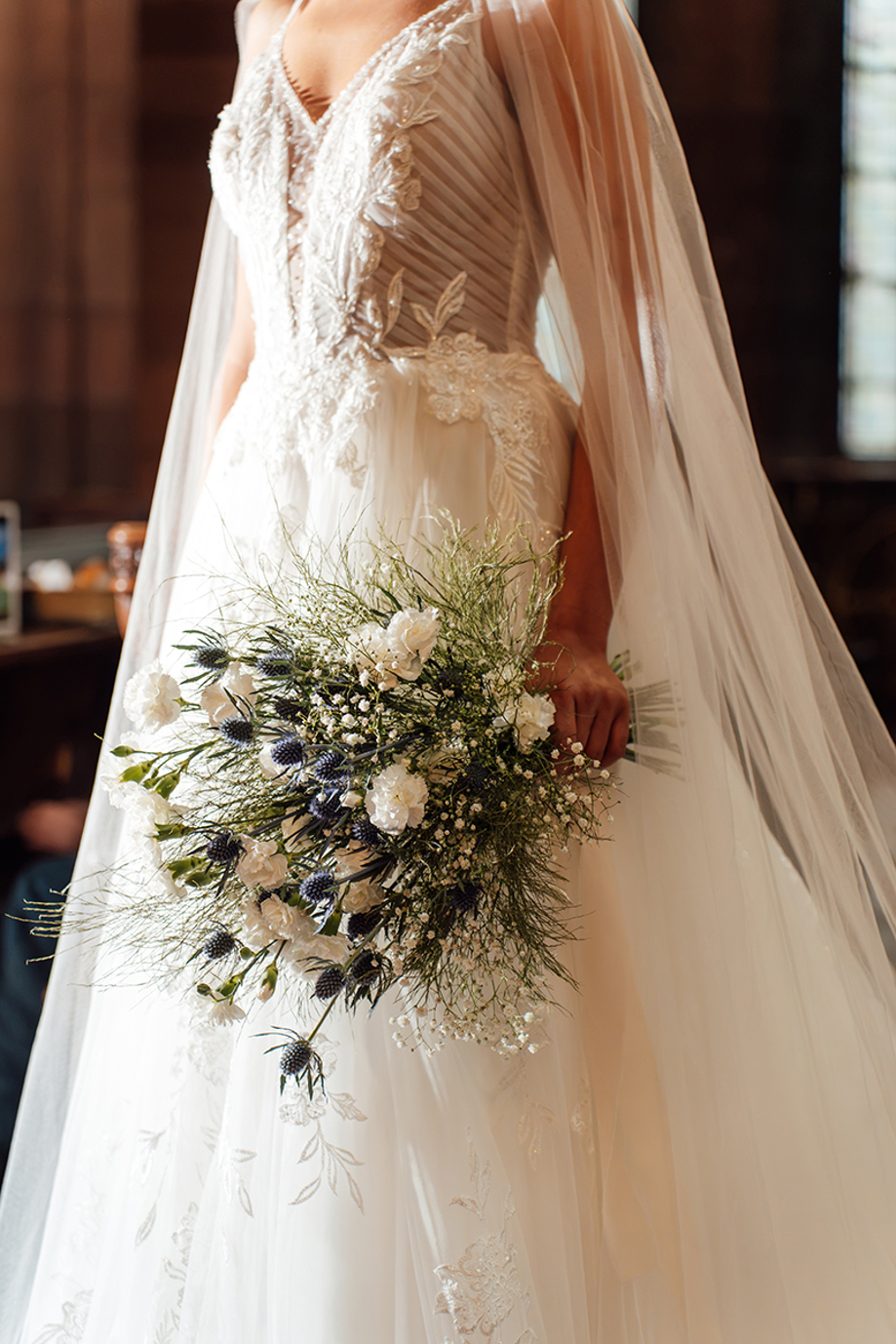 Close-up of a cascading bridal bouquet with white flowers, blue thistles, gypsophila and greenery by Maison Bloom Florals.