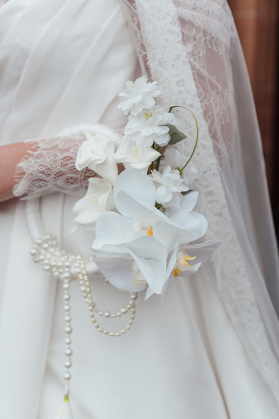 Close-up of white orchid and calla lily bridal flowers styled with pearl strands against a lace wedding dress.