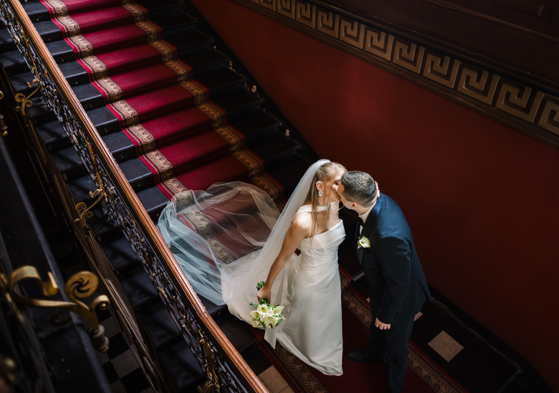 A bride in a white halter-neck gown with a veil and groom in a dark suit kissing on a grand staircase with a red runner and ornate black iron railing.