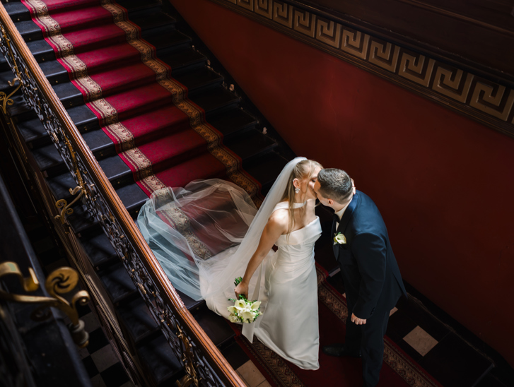 A bride in a white halter-neck gown with a veil and groom in a dark suit kissing on a grand staircase with a red runner and ornate black iron railing.