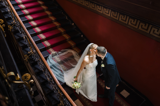 A bride in a white halter-neck gown with a veil and groom in a dark suit kissing on a grand staircase with a red runner and ornate black iron railing.