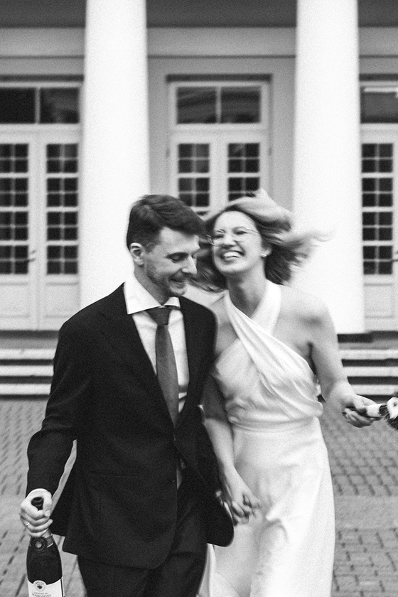 Black-and-white wedding photograph by Goda and Justas Photography of a newlywed couple laughing and running together, holding a bottle of champagne.