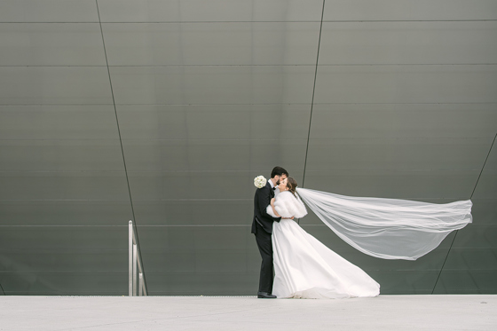 Bride in a fitted white wedding dress and long veil posing on an ornate staircase with red walls and decorative iron railings, photographed by Goda and Justas Photography.