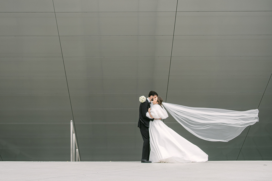 Bride in a fitted white wedding dress and long veil posing on an ornate staircase with red walls and decorative iron railings, photographed by Goda and Justas Photography.