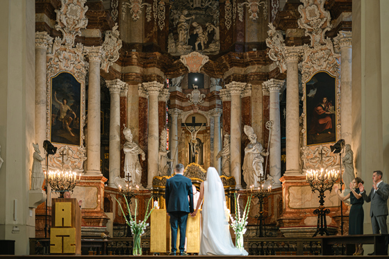 Catholic church wedding ceremony photographed by Goda and Justas Photography, showing a bride and groom holding hands at the altar beneath ornate religious sculptures and artwork.