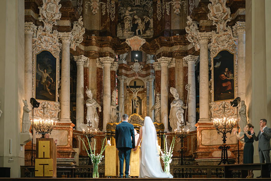 Catholic church wedding ceremony photographed by Goda and Justas Photography, showing a bride and groom holding hands at the altar beneath ornate religious sculptures and artwork.