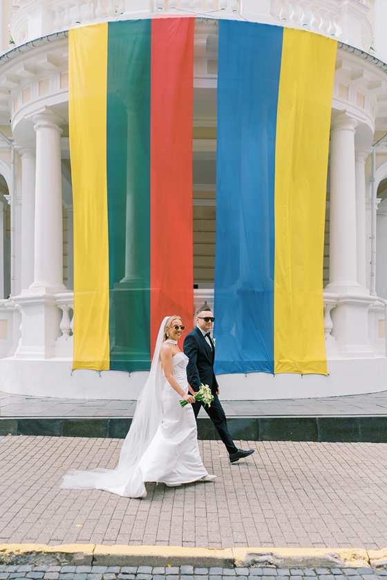 Bride and groom wearing sunglasses walking past a white building decorated with large colourful vertical fabric panels, photographed by Goda and Justas Photography.