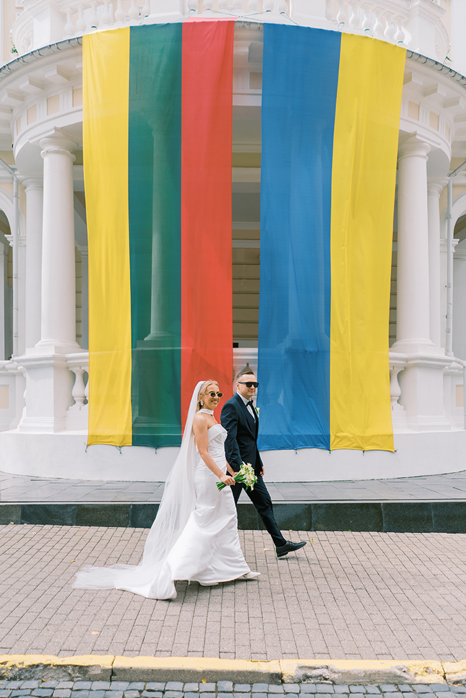Bride and groom wearing sunglasses walking past a white building decorated with large colourful vertical fabric panels, photographed by Goda and Justas Photography.