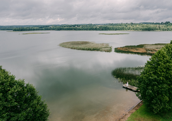 Aerial wedding photograph by Goda and Justas Photography showing a bride standing on a wooden jetty beside a large lake surrounded by reeds and woodland.
