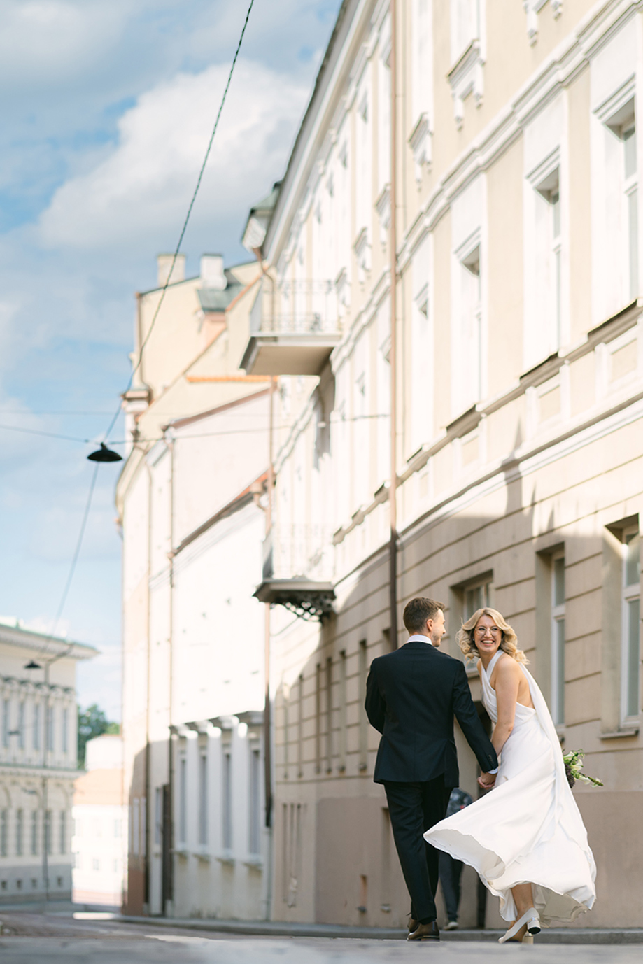 Candid wedding photograph by Goda and Justas Photography of newlyweds walking hand in hand along a city street, with the bride laughing in a flowing white halterneck dress.