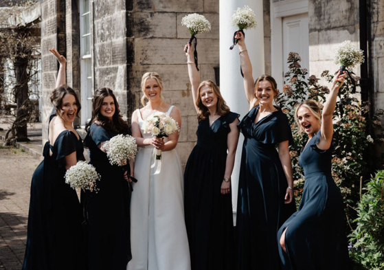Bride and bridal party in dark dresses posing with white bouquets outside a wedding venue, with bridal beauty by Bridal by Regina.