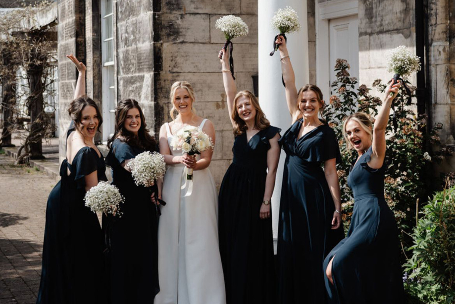 Bride and bridal party in dark dresses posing with white bouquets outside a wedding venue, with bridal beauty by Bridal by Regina.