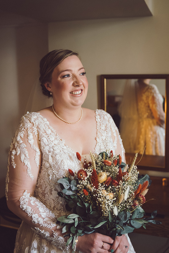 Bride in a lace wedding dress holding an autumnal wildflower bouquet with natural bridal makeup by Bridal by Regina.