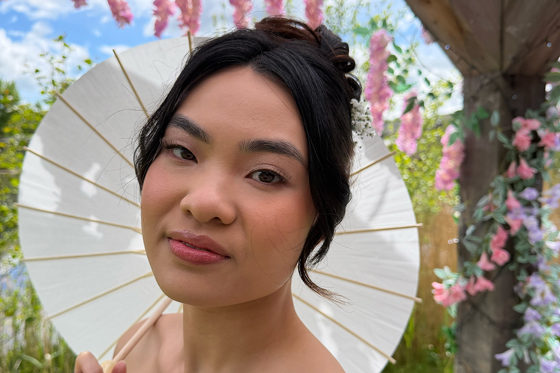 Bride holding a white parasol in a garden, showing soft pink bridal makeup and an elegant updo by Bridal by Regina.