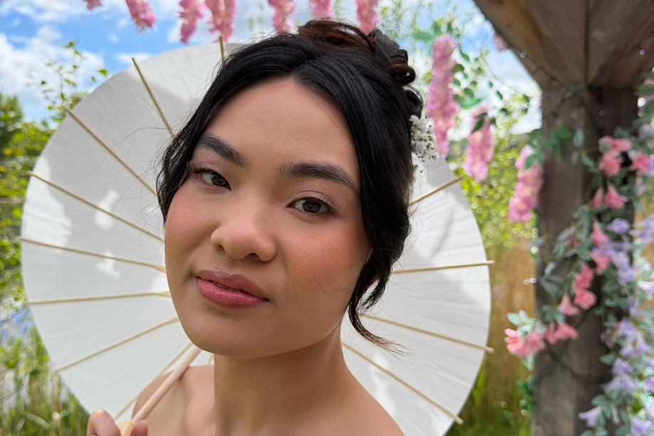 Bride holding a white parasol in a garden, showing soft pink bridal makeup and an elegant updo by Bridal by Regina.