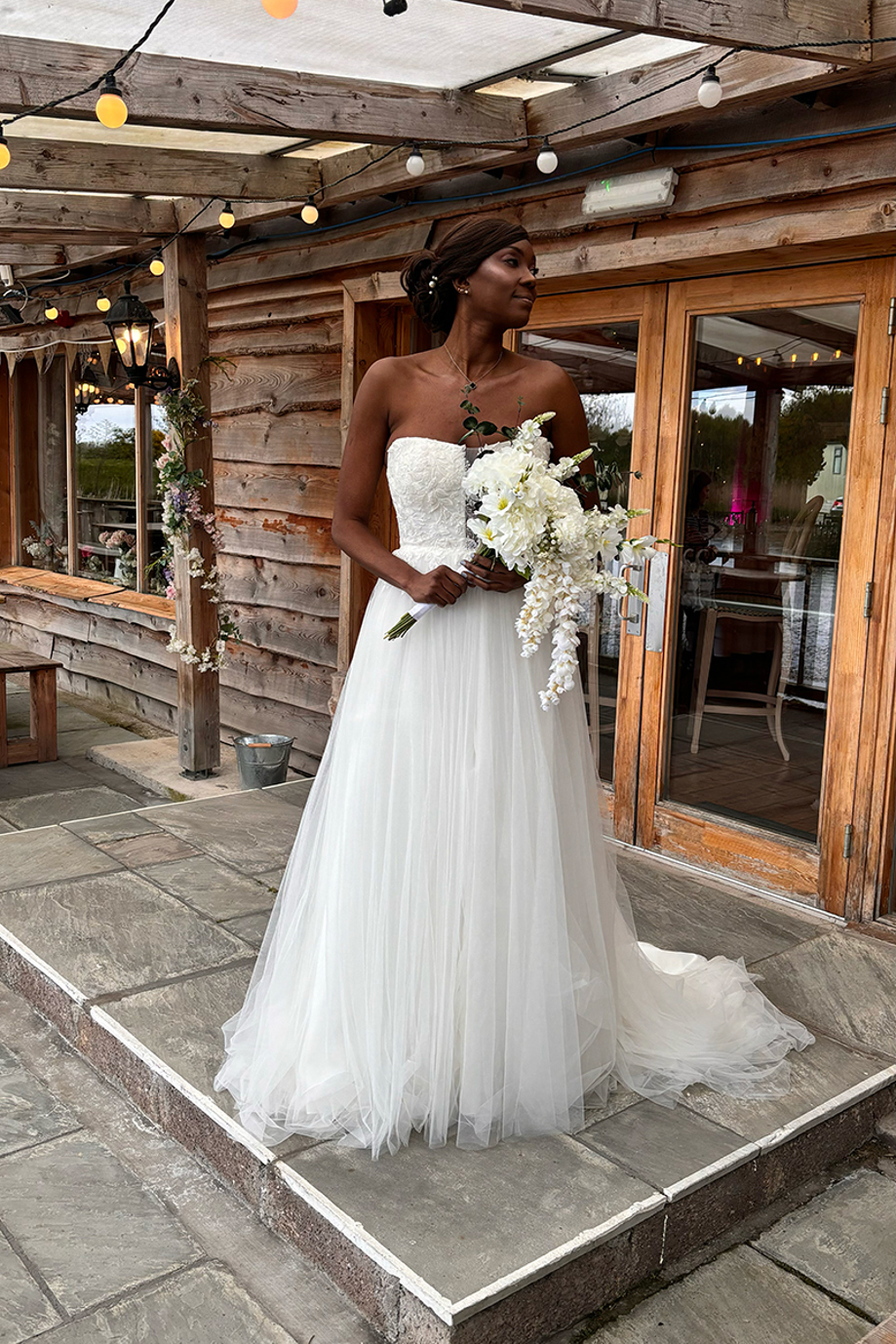 Bride in a strapless wedding dress holding cascading white flowers outside a rustic lodge, with bridal hair and makeup by Bridal by Regina.