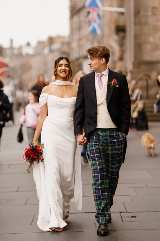 Newlywed couple walking through Edinburgh, with the bride wearing an off-the-shoulder wedding dress and elegant bridal hair and makeup.