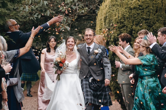 Newlyweds smiling as wedding guests throw confetti, with the bride wearing natural makeup and a relaxed bridal hairstyle.