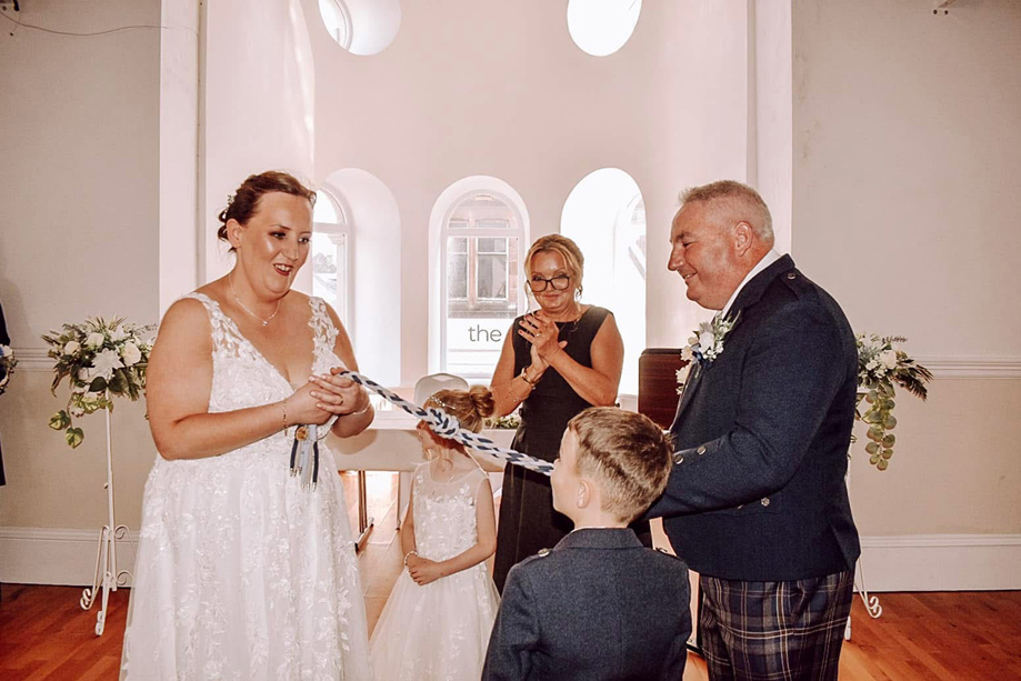 Bride and groom take part in a symbolic handfasting ceremony with their children and Fiona MacAlister Celebrant.