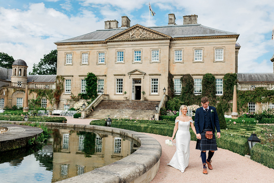 Bride and groom wearing traditional Scottish wedding attire walk beside a fountain at a country house wedding venue.