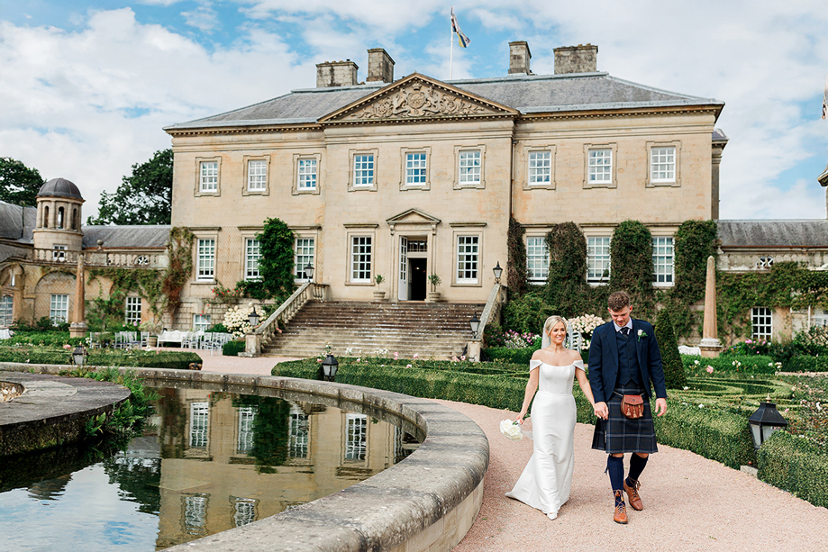 Bride and groom wearing traditional Scottish wedding attire walk beside a fountain at a country house wedding venue.