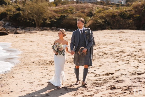 Bride and groom walk hand in hand along a Scottish beach, with the groom wearing a traditional tartan kilt outfit.