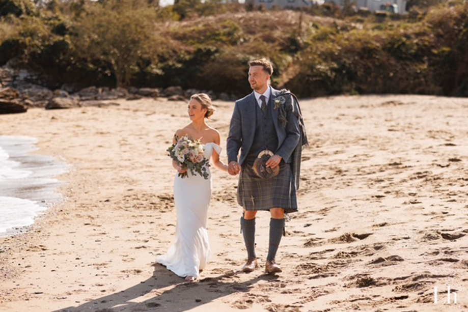 Bride and groom walk hand in hand along a Scottish beach, with the groom wearing a traditional tartan kilt outfit.