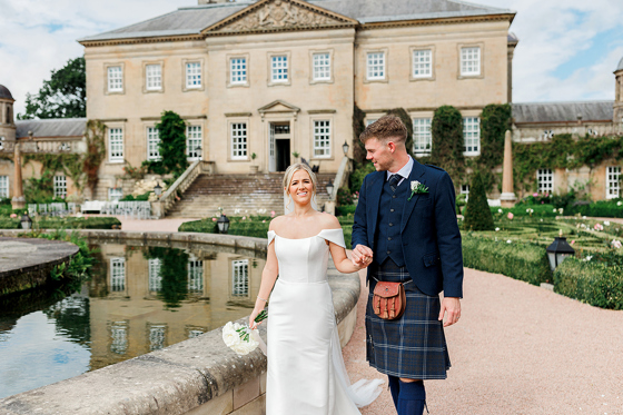 Bride in minimalist wedding dress and groom in navy tartan kilt walk around the gardens of a Scottish country house venue.
