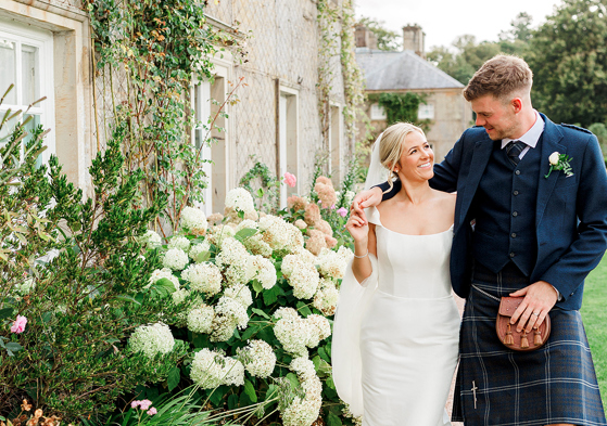 Newlyweds walk through flower-filled gardens at a Scottish wedding venue, with the groom wearing a traditional kilt outfit.