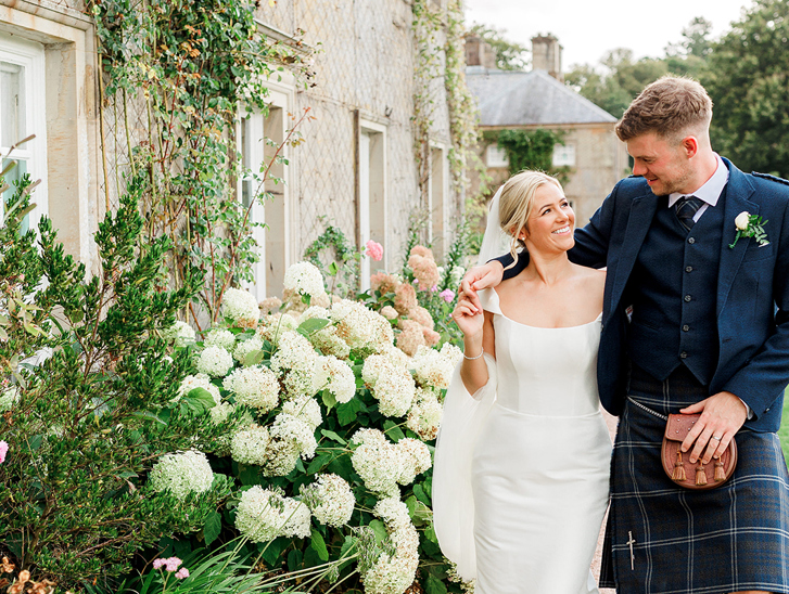 Newlyweds walk through flower-filled gardens at a Scottish wedding venue, with the groom wearing a traditional kilt outfit.
