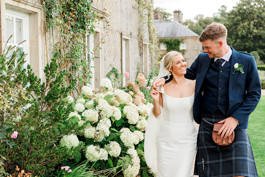 Newlyweds walk through flower-filled gardens at a Scottish wedding venue, with the groom wearing a traditional kilt outfit.