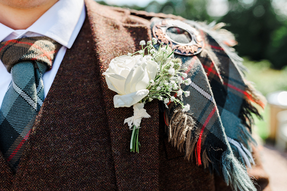 Close-up detail of groom’s Scottish wedding kilt, tartan sash and white floral buttonhole.