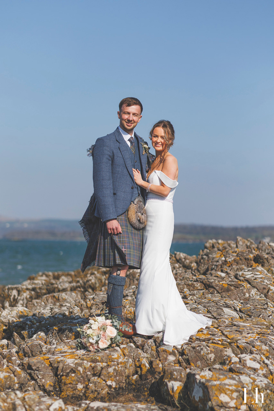 Bride and groom pose on coastal rocks overlooking the sea, showcasing traditional Scottish wedding kilt styling.