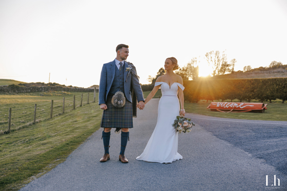 Bride and groom hold hands at sunset after their Scottish wedding, with the groom wearing a tartan kilt.