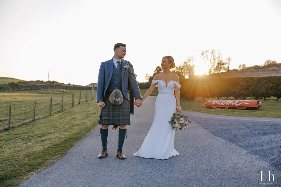 Bride and groom hold hands at sunset after their Scottish wedding, with the groom wearing a tartan kilt.
