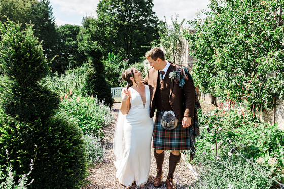 Bride and groom walk through colourful gardens at a Scottish wedding venue, featuring a traditional kilt wedding outfit.
