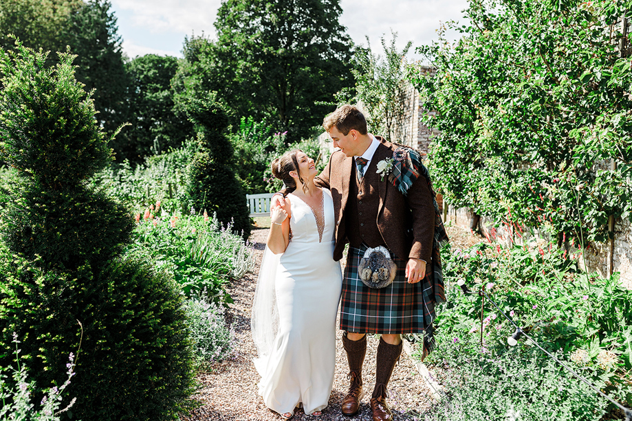 Bride and groom walk through colourful gardens at a Scottish wedding venue, featuring a traditional kilt wedding outfit.