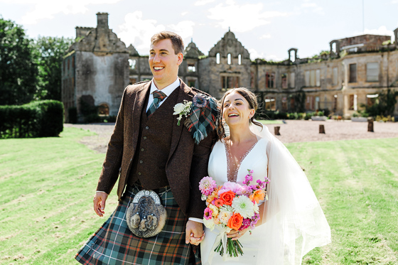Newlyweds laugh together outside a historic Scottish castle, with the groom wearing a brown tweed kilt outfit.
