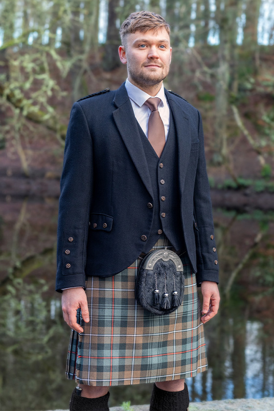 Groom stands in forest wearing kilt